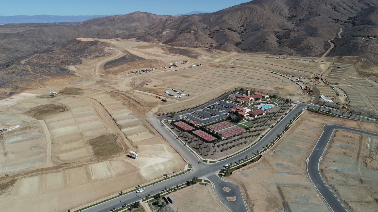 Aerial view of a clubhouse and pool, with tennis courts and open graded pads nearby